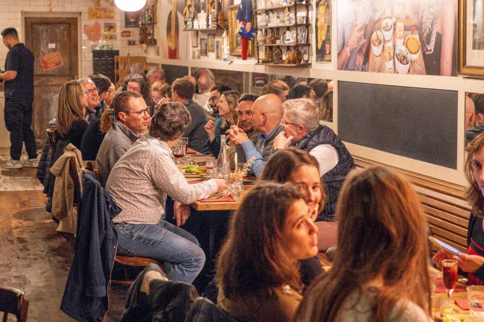 Eine lange Estaminet-Tafel mitten im Service, dicht sitzende Gäste, Schiefertafeln an der Wand.