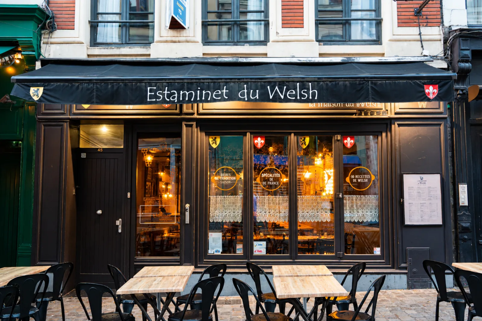 Die Fassade von L'Estaminet du Welsh, rue de Gand, schwarze Markise, beleuchtete Schaufenster und gedeckte Terrassentische auf dem Kopfsteinpflaster.