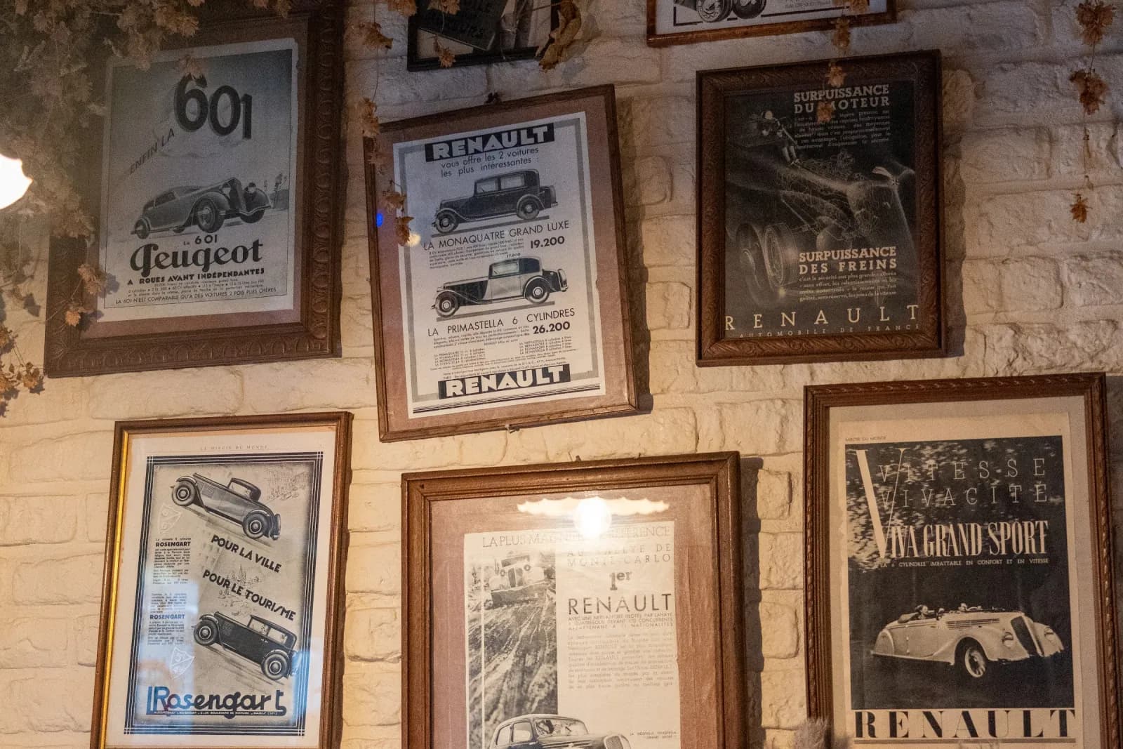 Un mur de brique blanchie couvert d'affiches automobiles encadrées, sous une guirlande de houblon séché.